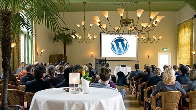 A meeting room with tables and attendees, looking at a large projector screen displaying the WordPress logo