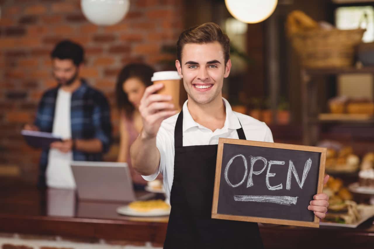 Portrait Of Smiling Barista Holding Take Away Cup And Open Sign At Coffee Shop