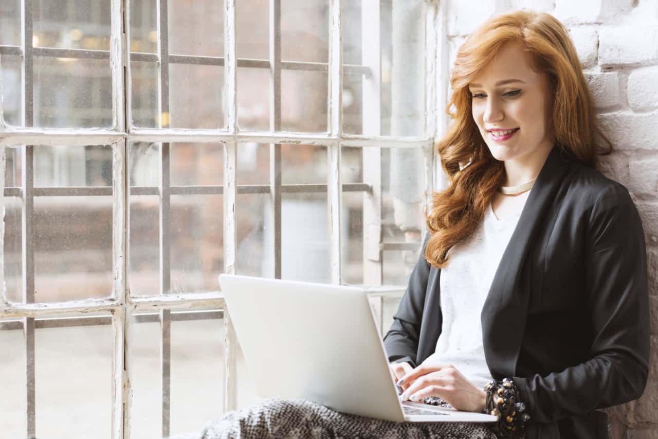 Woman Sitting Against Brick Wall Next To Window Typing On Laptop On Her Lap